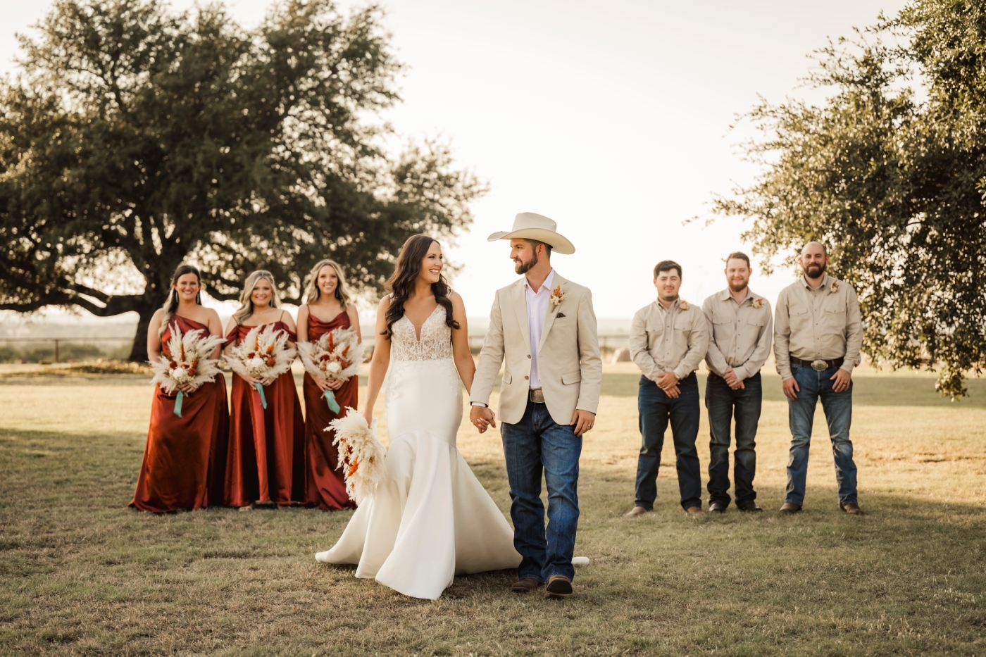 A wedding party with bridesmaids in silk, rust-colored dresses and groomsmen in tan smile at a bride and groom at a micro wedding at Diamond A Ranch