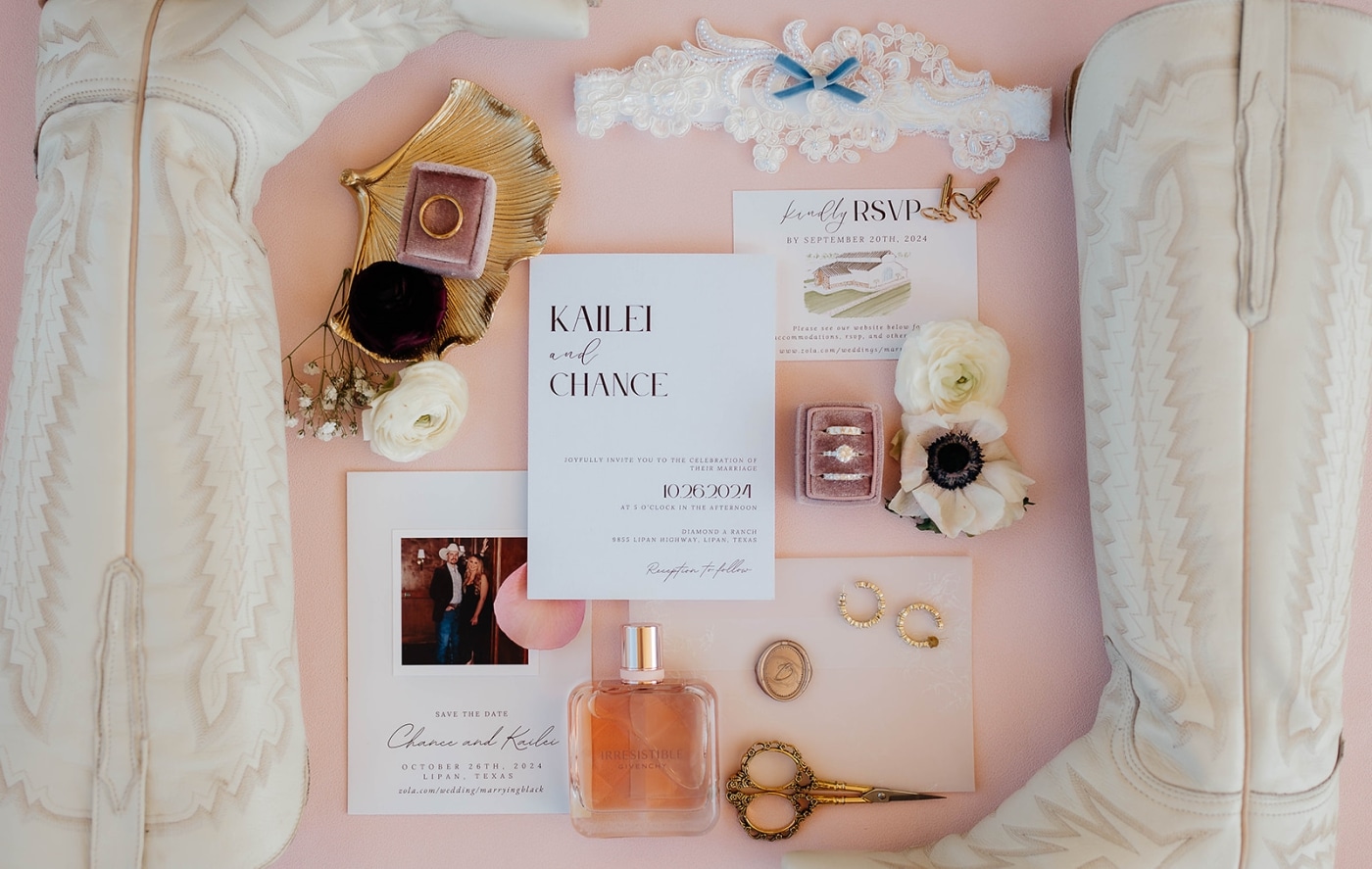 A flat lay of wedding invitations, white cowboy boots, and a bottle of perfume