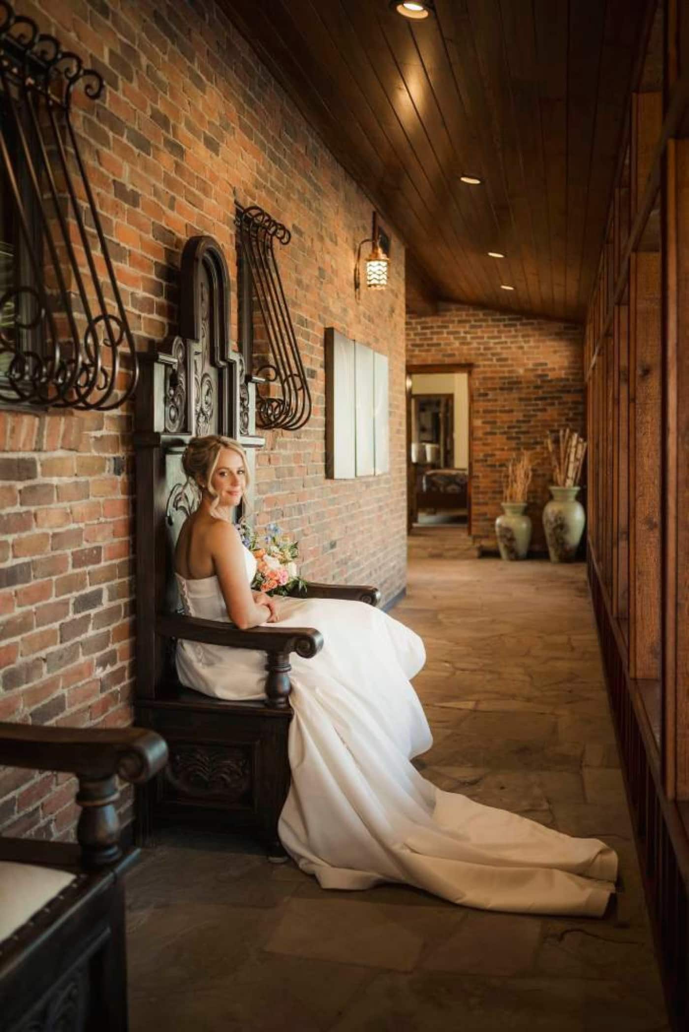 A bride sits on a carvebd wooden chair in a brick hallway