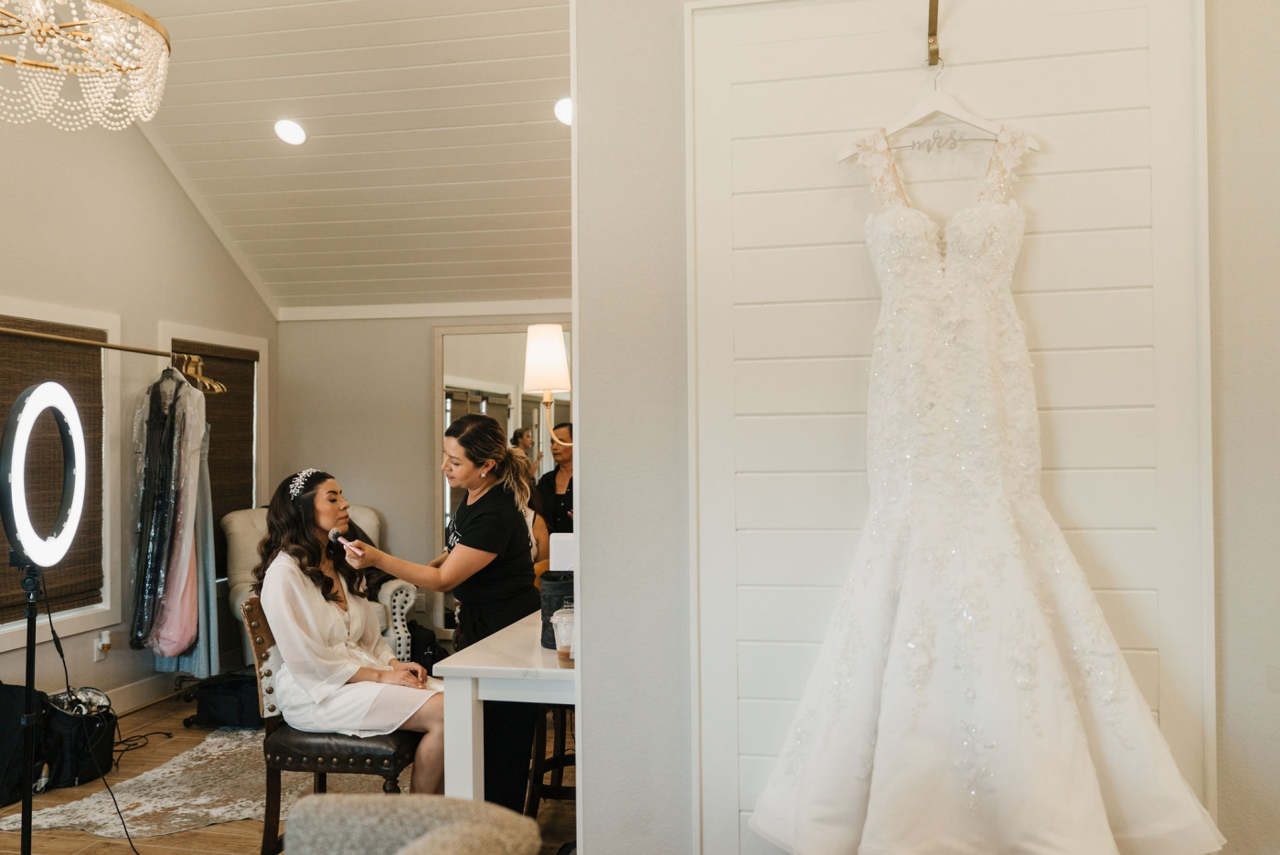A bride gets her makeup done while her wedding dress hangs on a hanger that says, "bride"